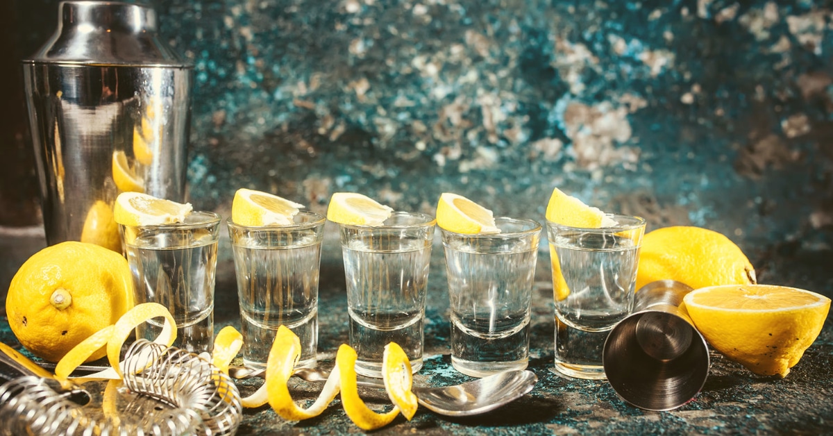five gin shots on a table with lemons, lemon twists, and a cocktail shaker