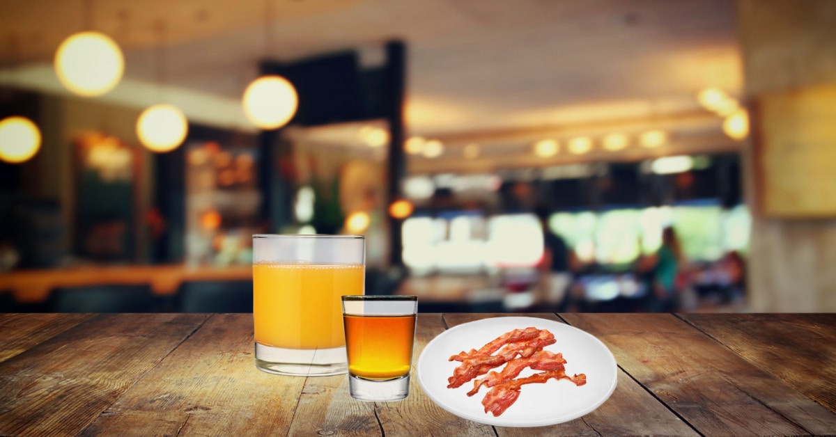irish breakfast shot recipe pictured with an irish whiskey and butterscotch liquor shot, a glass of orange juice, and a plate of bacon in a restaurant setting