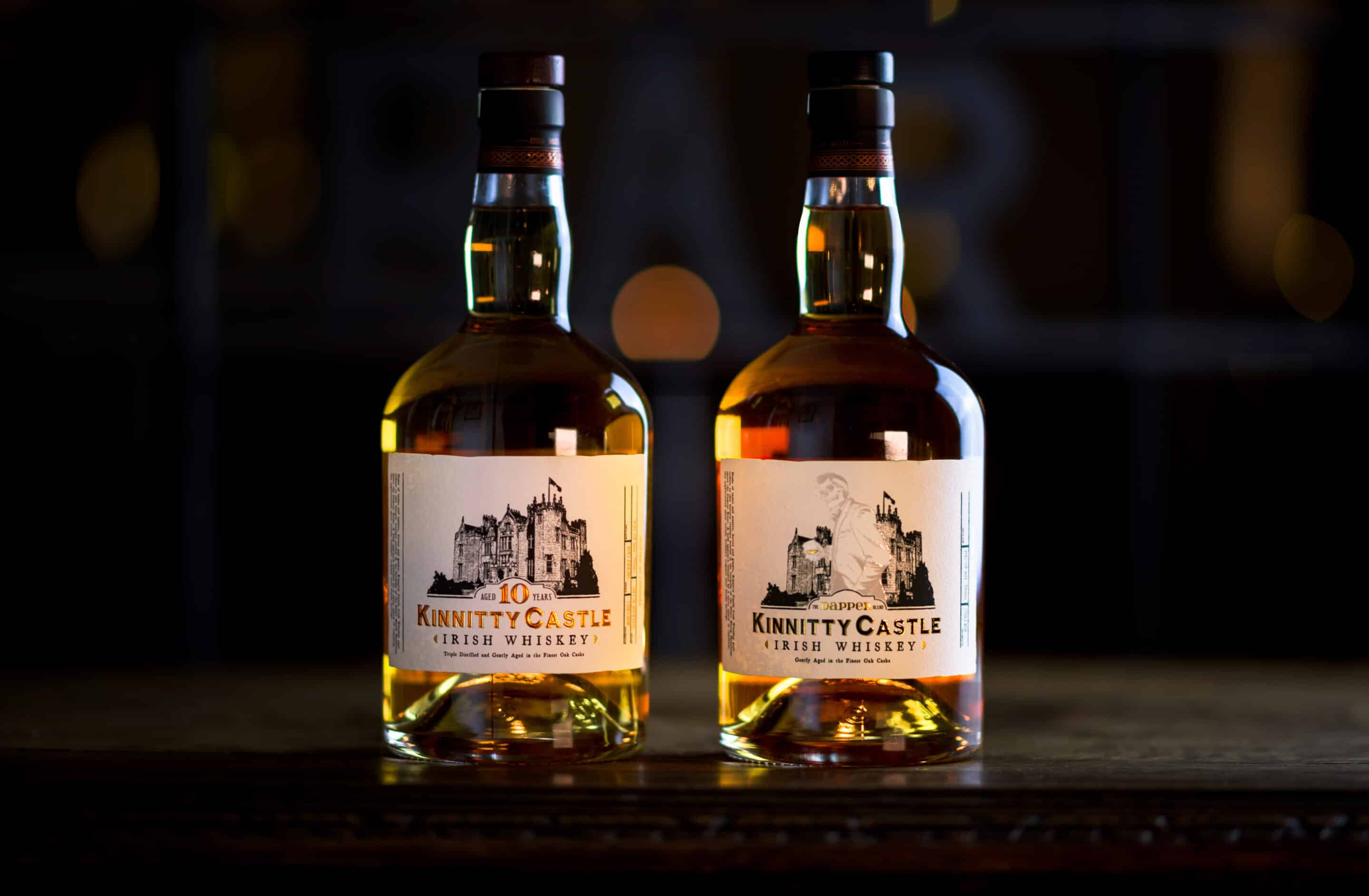 A bottle of Kinnitty Castle Irish Whiskey Aged 10 Years next to a bottle of Kinnitty Castle IRish Whiskey Dapper Blend on a table with a blurred background